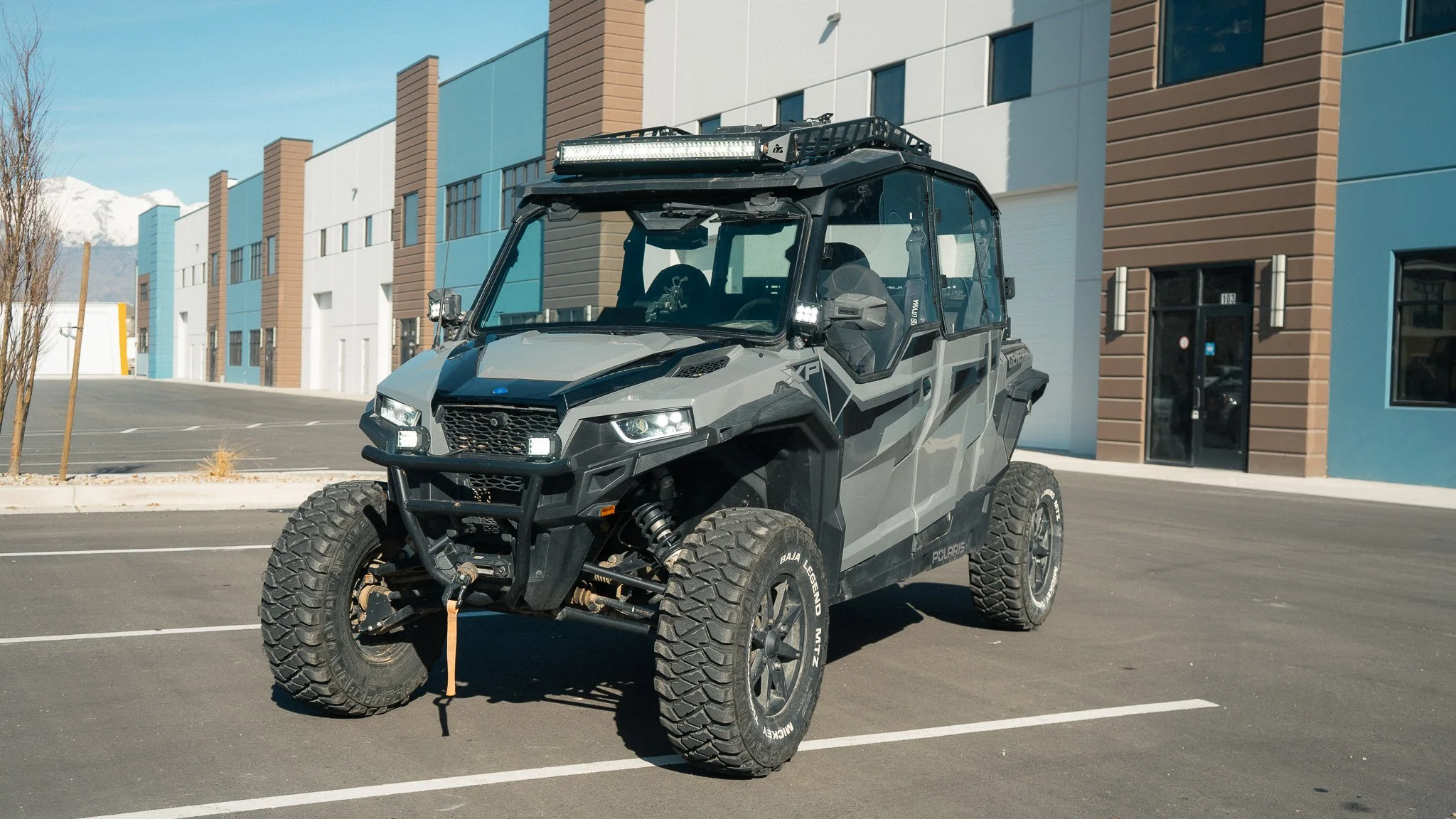 Black and gray off-road utility vehicle parked in an empty parking lot with modern multicolored buildings in the background.