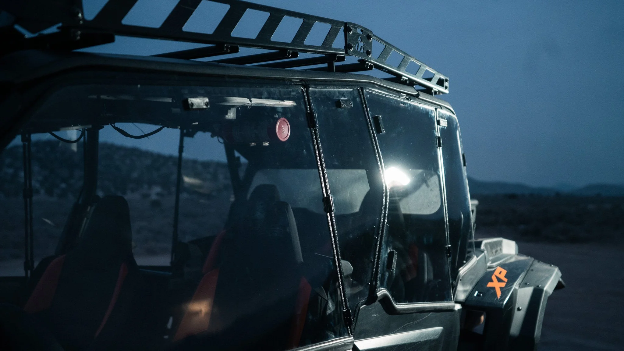 An off-road utility vehicle with a glass enclosure, parked in a desert landscape during dusk. The vehicle has a roof rack and orange accents, with the letters "XP" visible on the side.