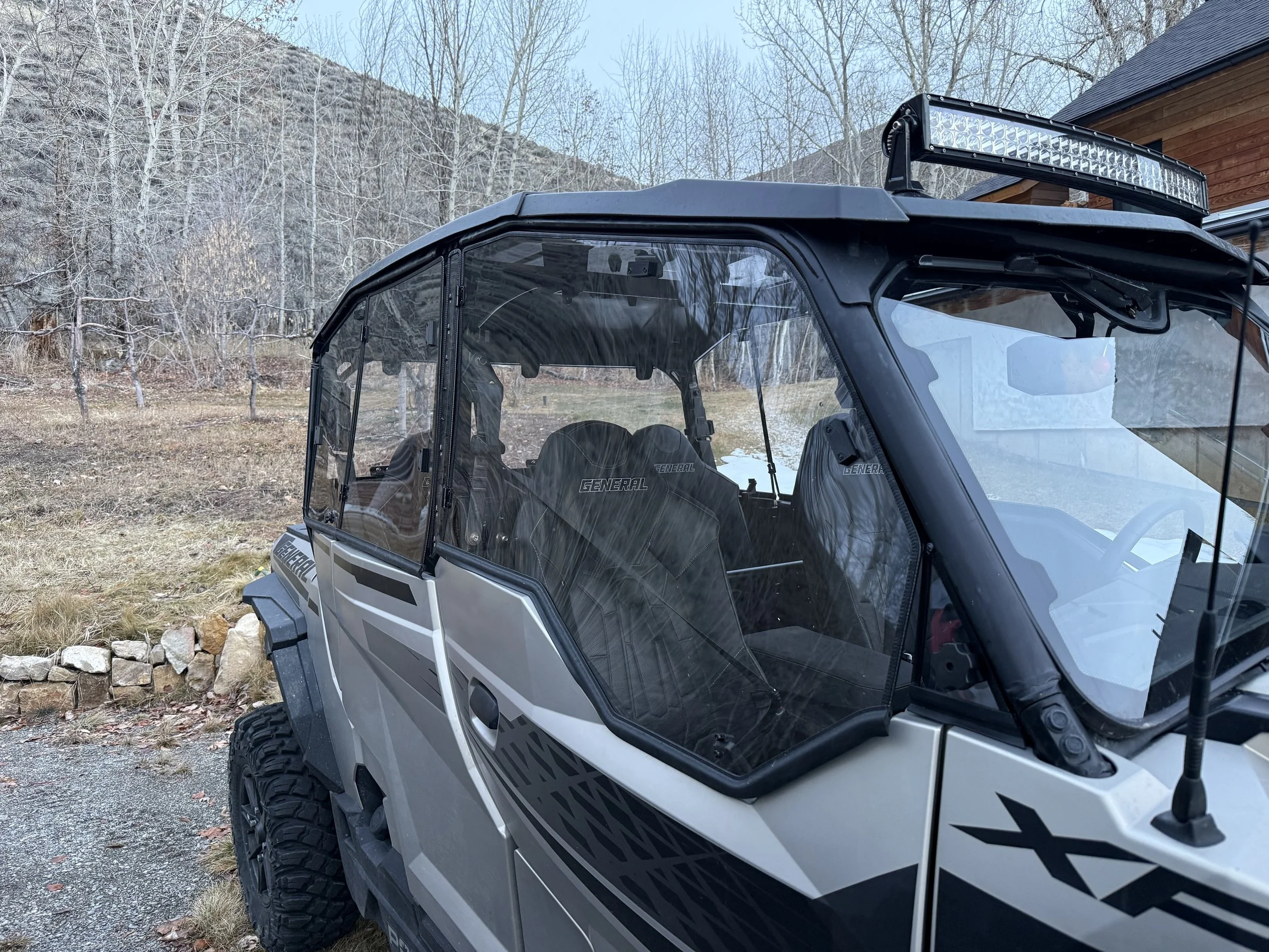Close-up of a black and silver off-road utility vehicle parked outdoors, with a mountain, leafless trees, and a building in the background.