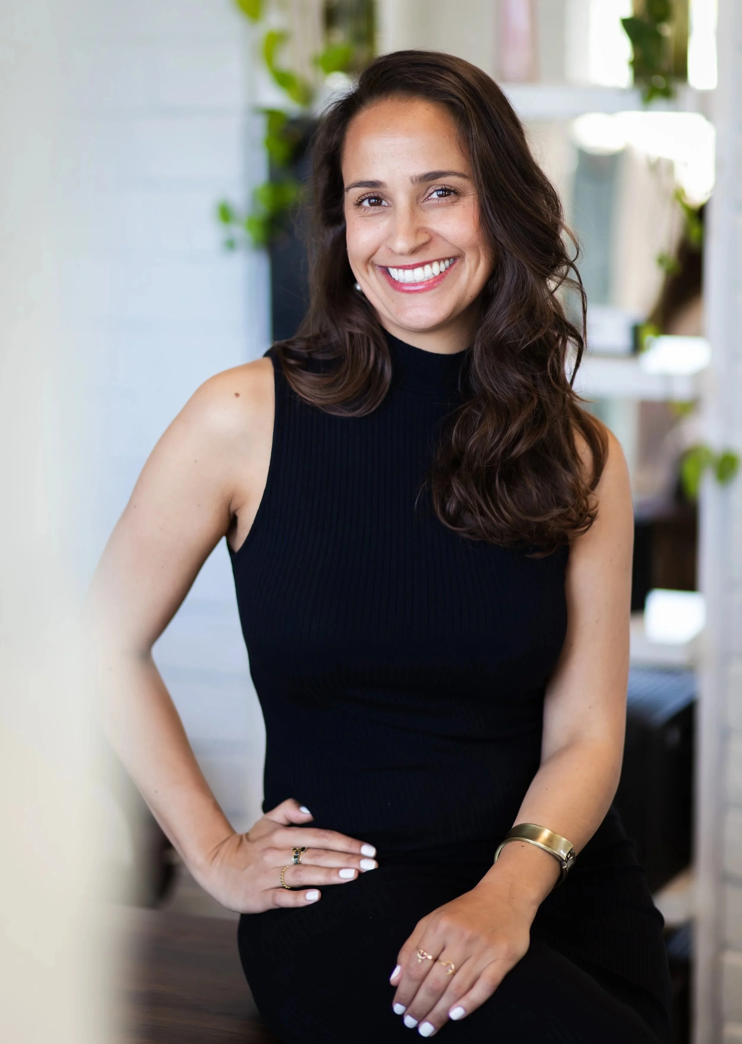 A woman with long, wavy brown hair smiling, wearing a sleeveless black top, sitting with her right hand on her hip, indoors with plants and shelves in the background.