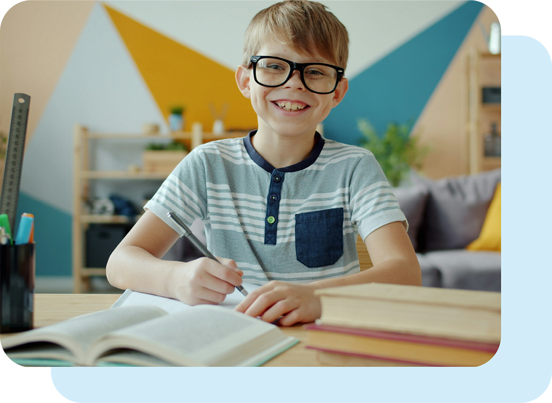 Smiling boy in glasses holds a pen in one hand as he gazes up from the books open in front of him on his desk