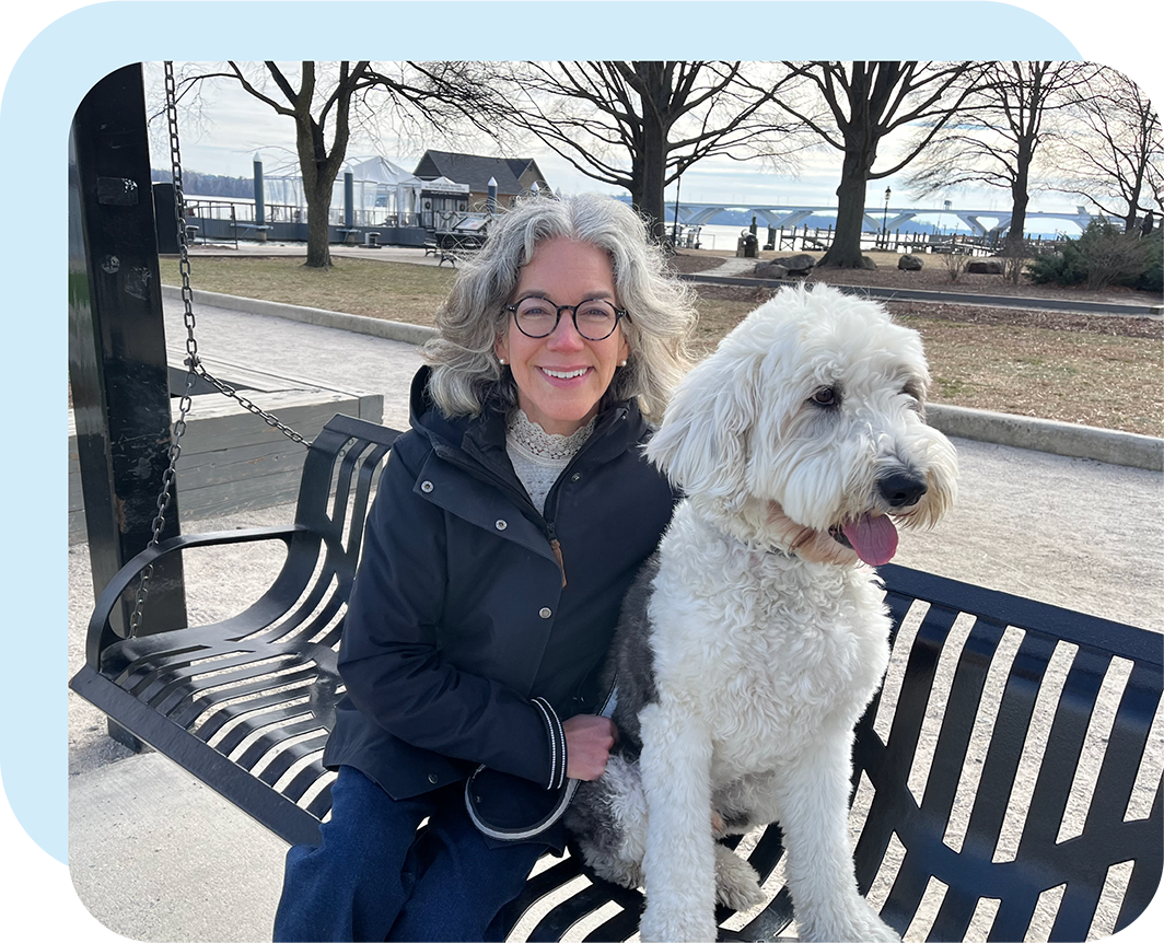Kristen Corwin, founder of Old Town Tutoring, sitting on a swing bench by the marina in Old Town Alexandria with her sheepadoodle, Watson, beside her, tongue out.