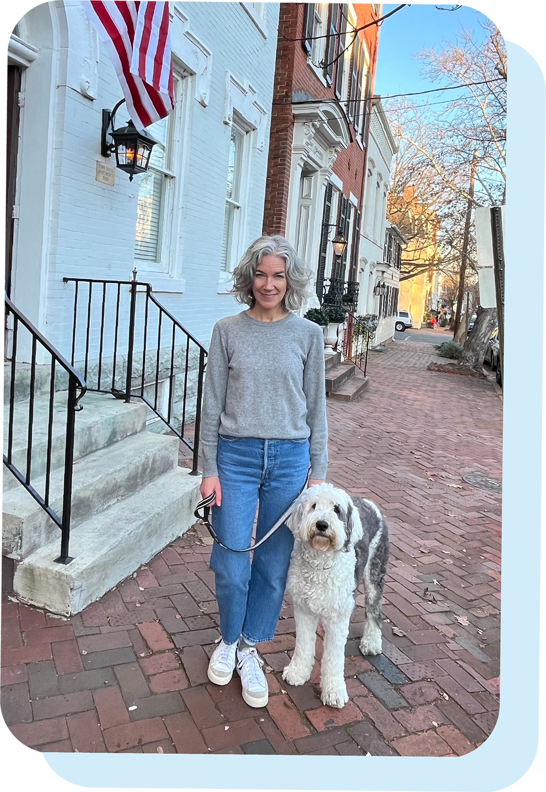 Kristen Corwin, founder of Old Town Tutoring, stands on a brick street in Old Town Alexandria wearing a grey sweater, blue jeans, and white sneakers. Her white and grey sheepadoodle, Watson, stands beside her.
