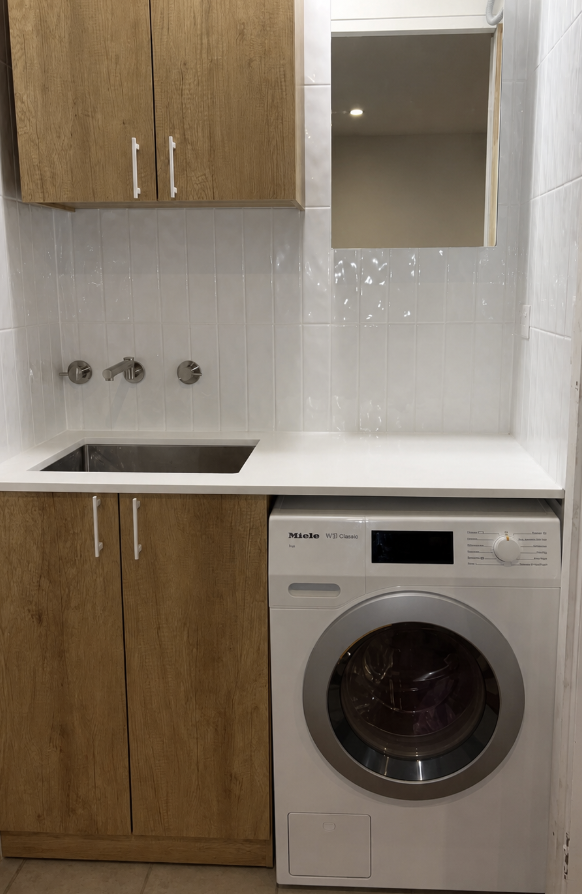 Laundry room with wooden cabinets above and below a white countertop. Stainless steel sink with wall-mounted faucets. Mirror above the countertop. Front-loading washing machine below the right side of the countertop.