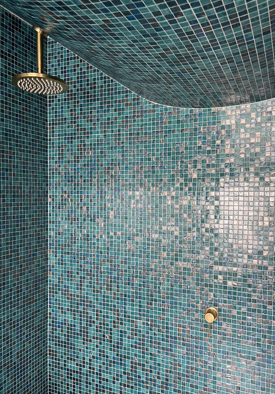 Interior of a shower room with turquoise mosaic tiles, a round gold showerhead from the ceiling, and a small gold control knob on the tiled wall.