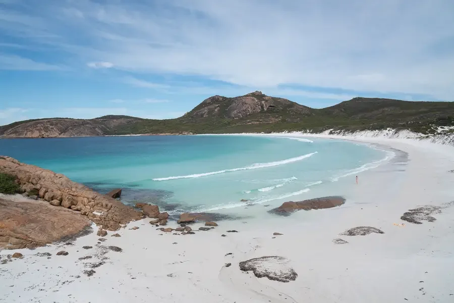 A pristine beach with white sand, turquoise ocean waves, rocky formations, and green hills with a mountain in the background under a partly cloudy sky.
