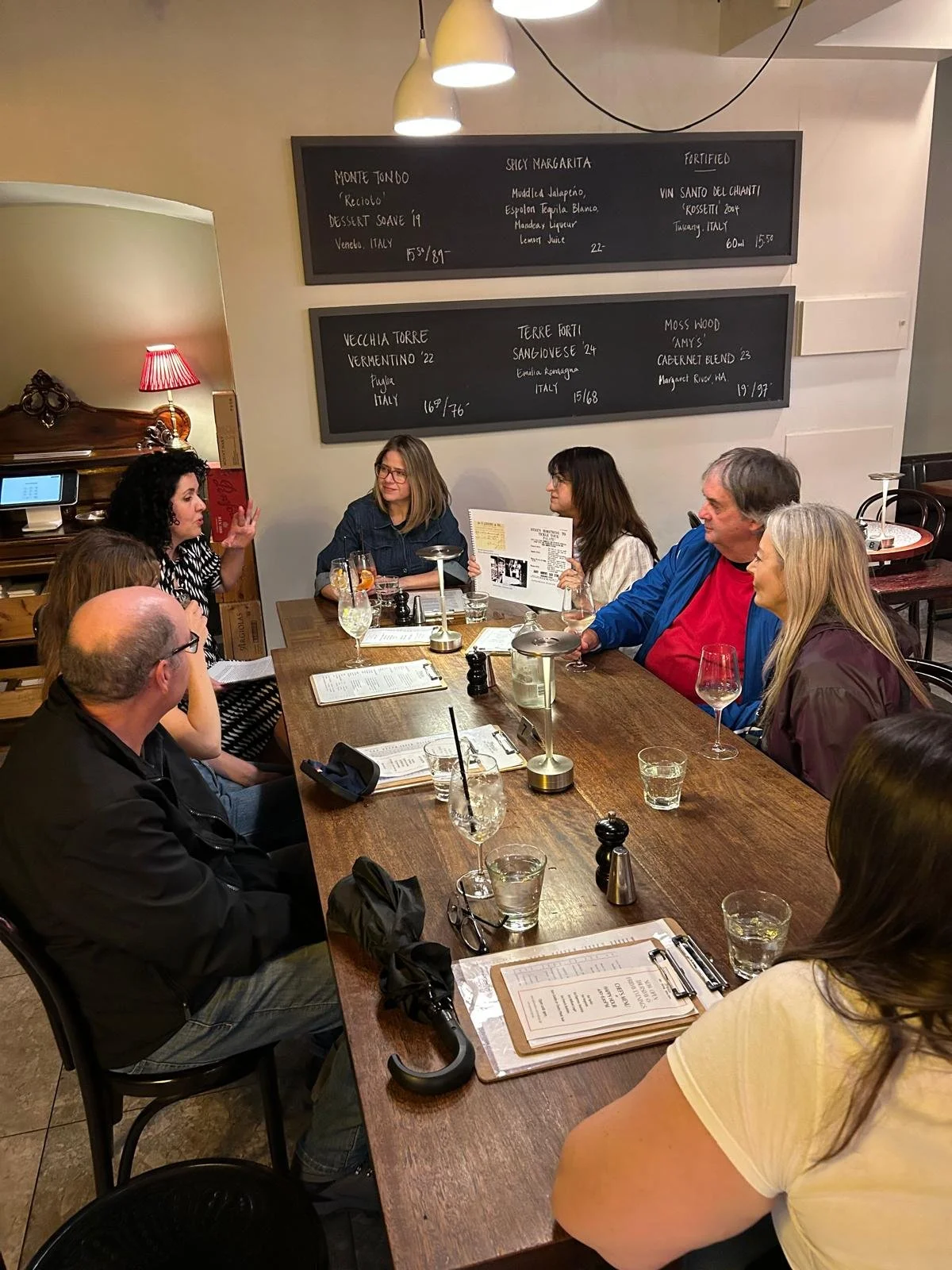 A group of people sitting around a long wooden table in a restaurant, with black chalkboard menus on the wall behind them and drinks on the table.