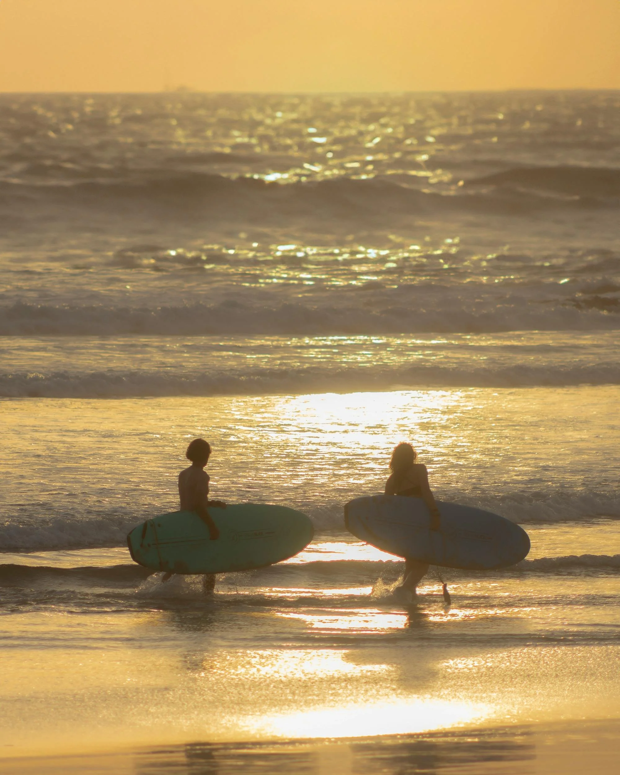 Two people with surfboards walking into the ocean during sunset.