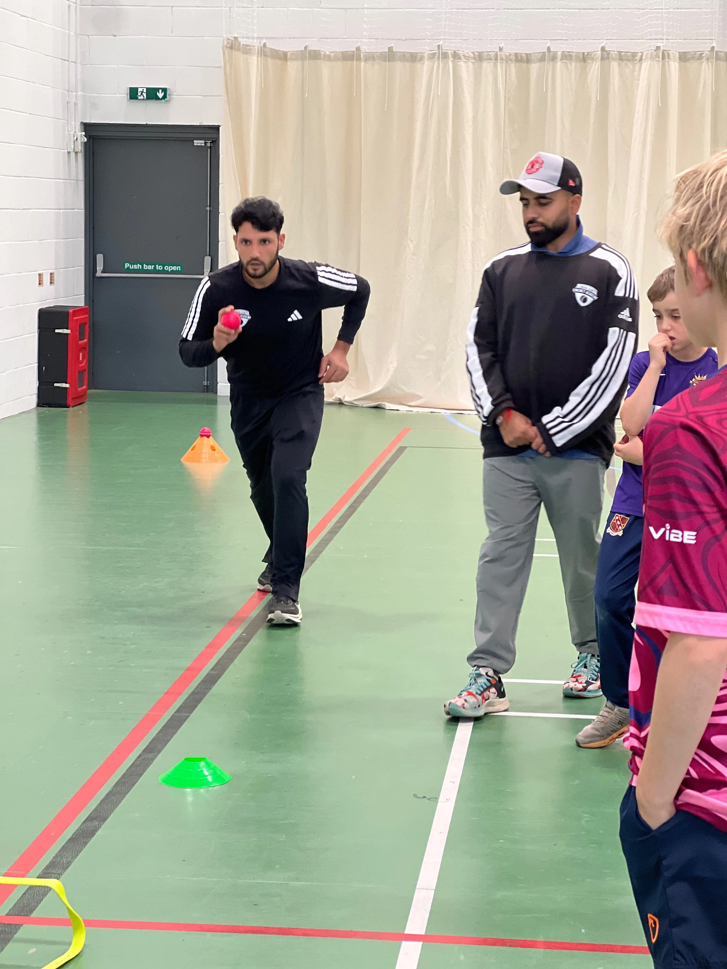 A man in black tracksuit throwing a pink ball in an indoor sports hall, with other children and a coach watching.