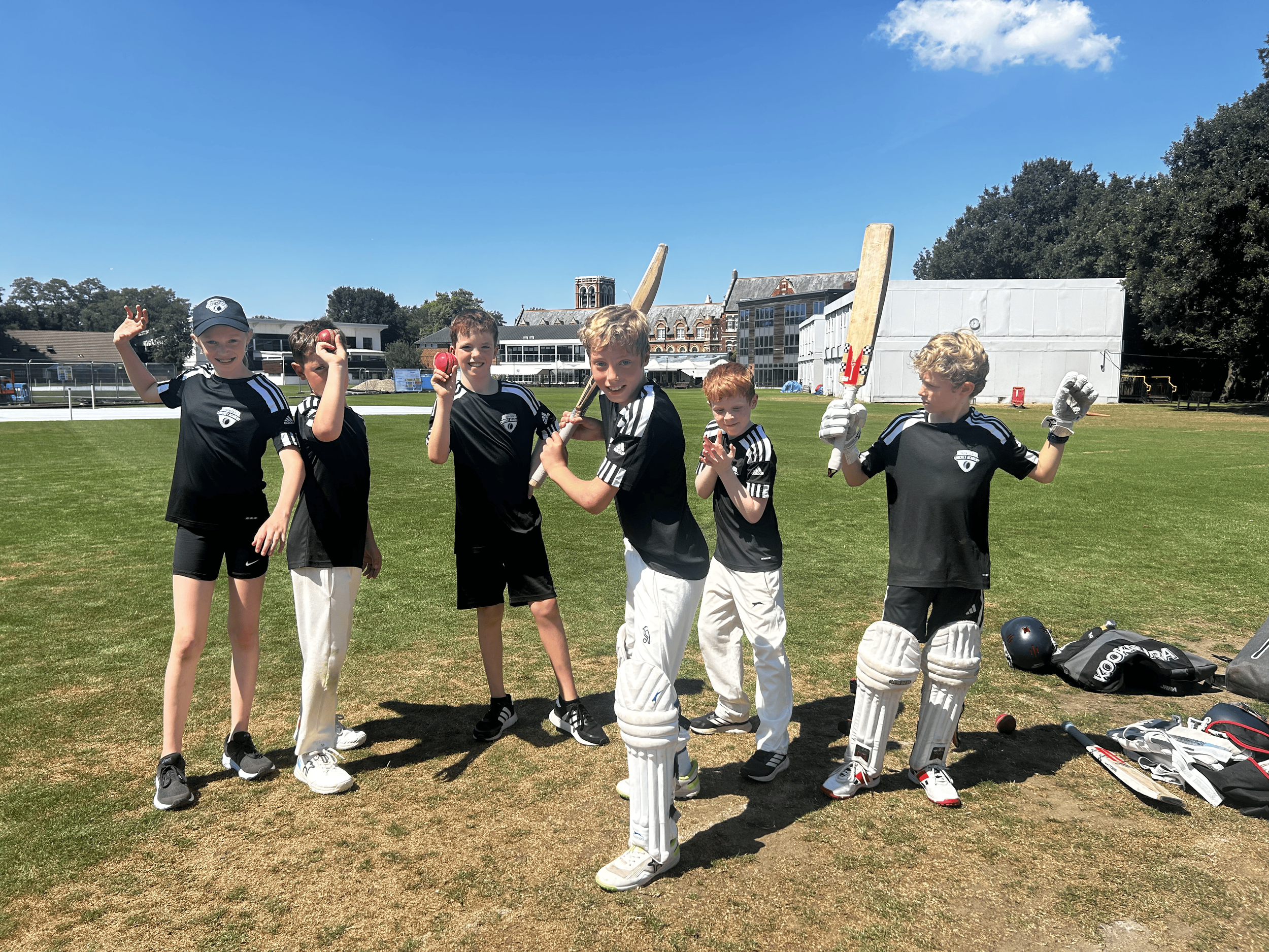 Six young boys in cricket uniforms pose on a cricket field with cricket pads and bats, smiling and showing various hand gestures under a clear blue sky.