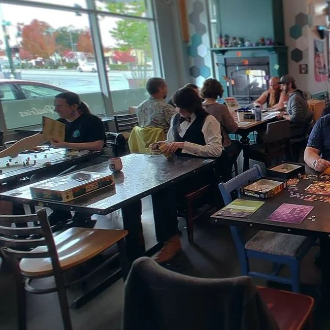 People playing board games and reading in a cozy cafe or game spot with large windows and colorful wall decor.