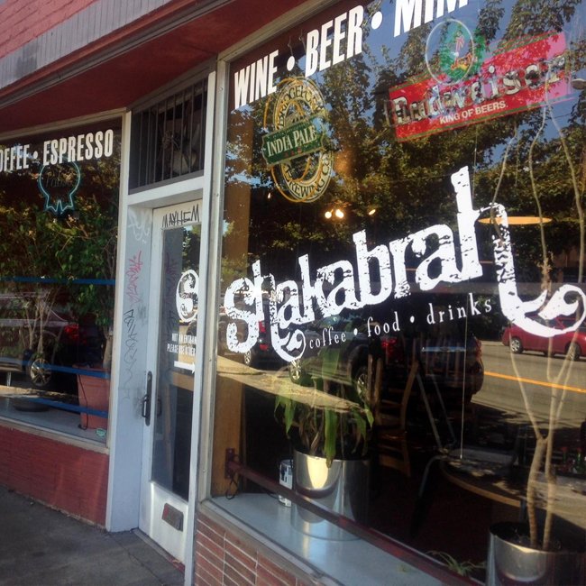 Exterior view of a café named Sakahabi with large glass windows displaying stickers for wine, beer, and coffee. The window also has text for coffee, food, and drinks, and the door has a small sign and some graffiti. Inside, potted plants are visible.