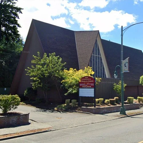 Exterior view of a modern church building with wooden triangular architecture, large glass windows, and trees and bushes in front.
