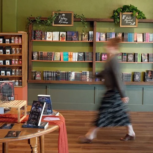 A woman walking past bookshelves filled with colorful books in a bookstore or library, with wooden flooring and green walls, and signs smiling with decorative greenery on top.