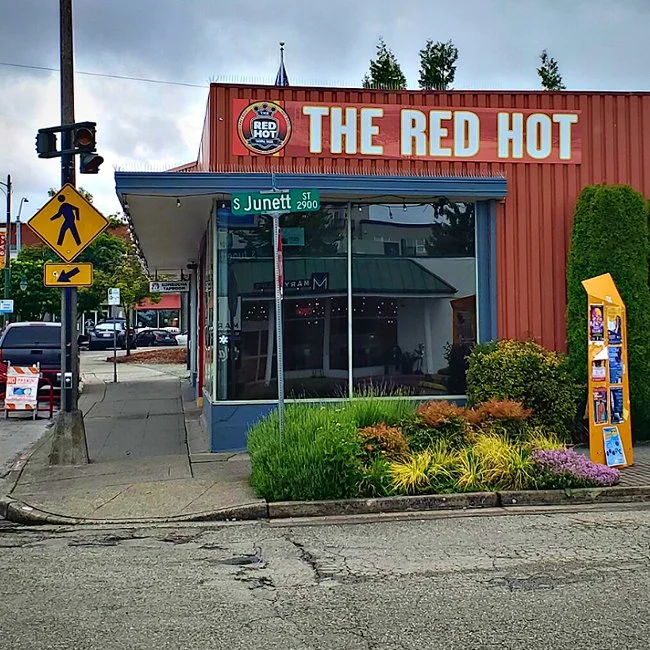 Street view of a building named 'The Red Hot' with a shop window, a street sign 'S Junett St 2900', a yellow pedestrian crossing sign, and a yellow parking meter outside. There are plants and bushes in front of the building.