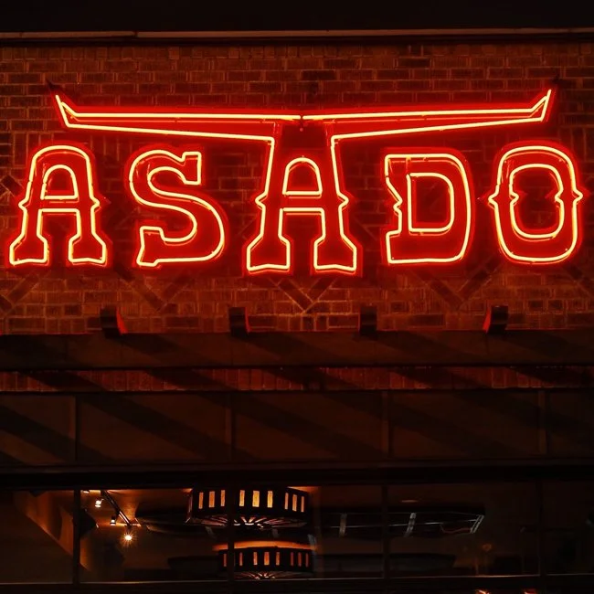 Neon sign on brick wall displaying the word 'ASADO' with a decorative line above it.