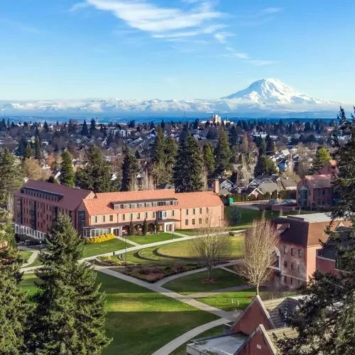 A scenic view of a suburban neighborhood with University of Puget Sound buildings and green lawns in the foreground, and Mount Rainier in the distance under a partly cloudy blue sky.
