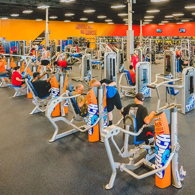 People working out on various gym equipment in a spacious fitness center with bright yellow and orange walls and multiple TV screens.