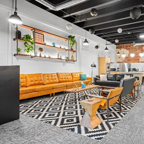 Modern lounge area with a brown leather sofa, black armchair, wooden coffee table, and geometric side table, decorated with hanging and wall plants, in an industrial-style space with a black ceiling, exposed ductwork, and decorative paper lanterns.