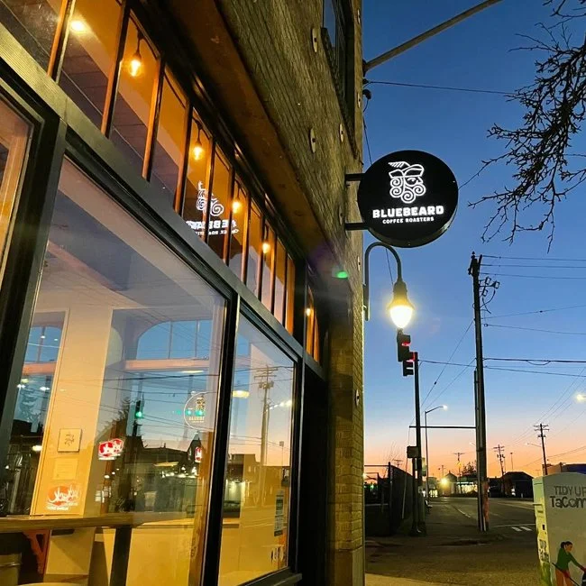 Exterior of Bluebeard Coffee Roasters storefront at dusk, with a black round sign featuring a white stylized skull logo, large glass windows showing the interior, and streetlights illuminating the sidewalk.