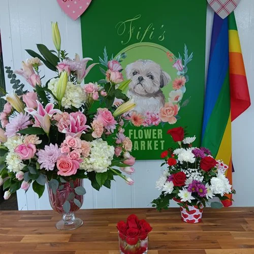 Various floral arrangements including pink roses, lilies, and mixed flowers in vases on a wooden table, with a green sign and colorful flags in the background.