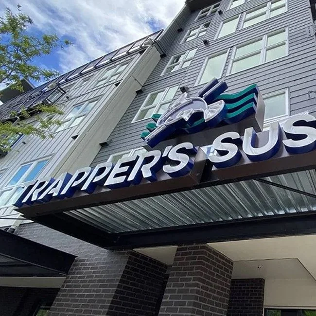 Sign for Trapper's USU in front of a multi-story building with gray siding and multiple windows, under a partly cloudy sky.