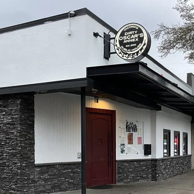 Exterior of a building housing 'Dirty Oscar's Annex', with a round sign on the corner, a maroon door, and a white wall with a black logo and gray stone accents.