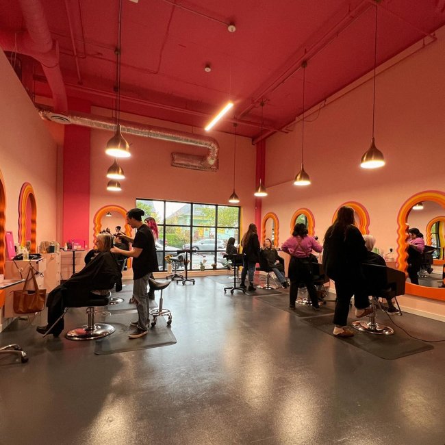 Inside a colorful hair salon with pink walls, large mirrors, and multiple clients getting hair treatments while stylists work. Natural light from large windows illuminates the space.