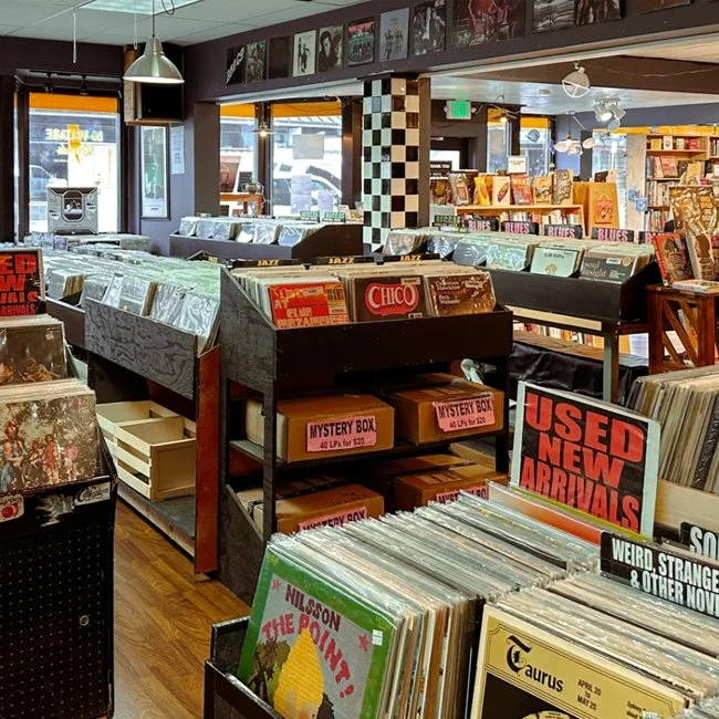 Interior of a record store with shelves of vinyl albums, display boxes labeled 'Mystery Box' and signs advertising used, new arrivals, and various music genres; posters and photographs on the walls.