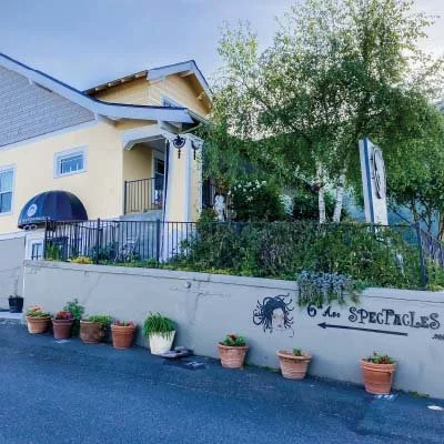 A yellow two-story building with a blue roof, a small balcony, and potted plants outside. A wall with girl graffiti and the words "6 underground" is in front of the building, along a street with a black fence and trees.