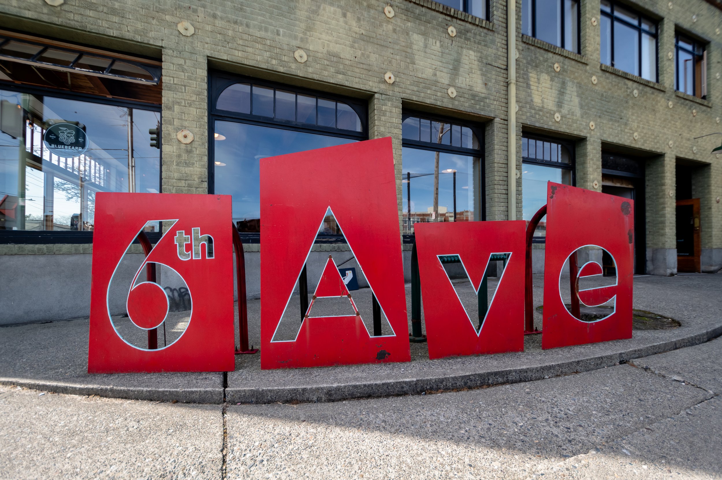 Red sculpture spelling '6th Ave' in front of a brick building with large windows.