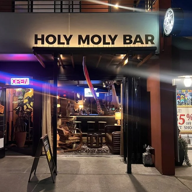 Entrance to Holy Moly Bar with illuminated sign, open neon sign, and visible interior with seating and bar area.