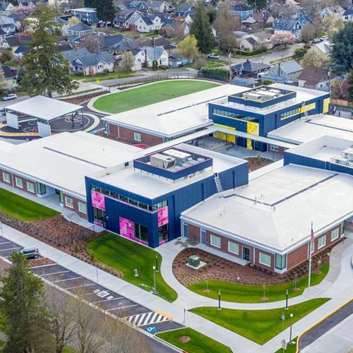 An aerial view of a school with multiple flat-roofed buildings, a large outdoor sports field, and surrounding residential neighborhood.