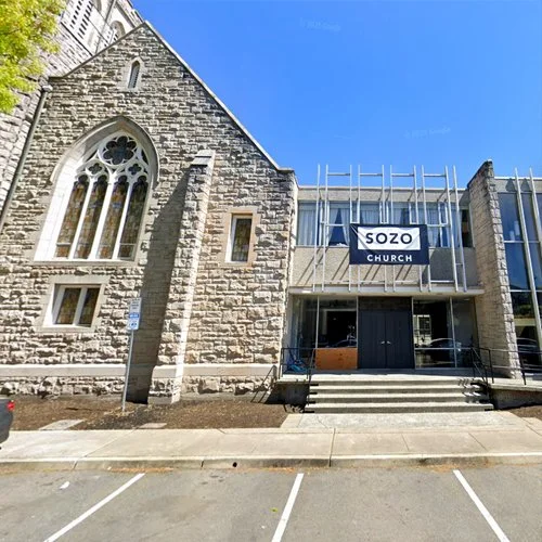 A stone church building with arched windows on the left and a modern addition under construction on the right, with a sign reading 'SOZO CHURCH' on the scaffolding.