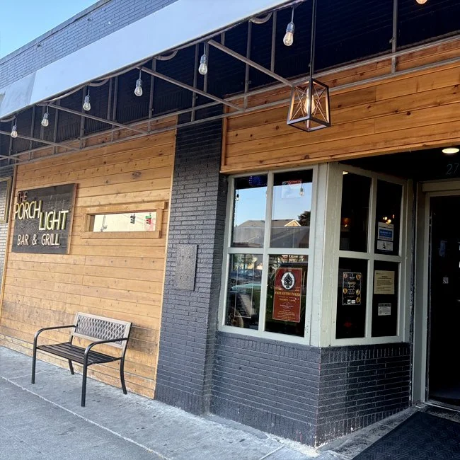 Exterior view of a restaurant named "The Porch Light Bar & Grill," showing a wooden facade with a bench outside, large windows, and string lights hanging above.