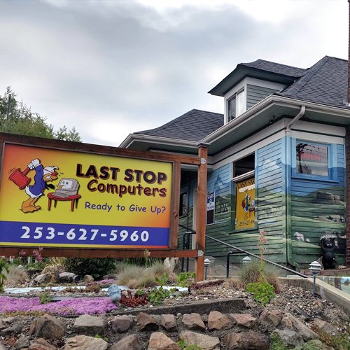 Sign for Last Stop Computers featuring a cartoon bird with a cape and a computer, with phone number 253-627-5960, in front of a blue house with murals and posters.