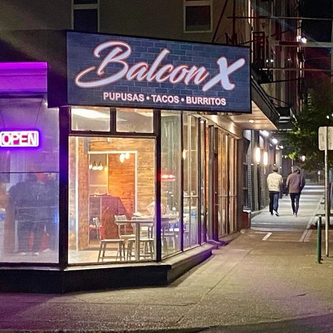 Nighttime view of a restaurant called Balcon X with a sign advertising pupusas, tacos, and burritos. The interior is visible through large glass windows, showing tables and chairs, with some indoor lighting. An illuminated OPEN sign is on the left, and two people are walking on the sidewalk past the restaurant.
