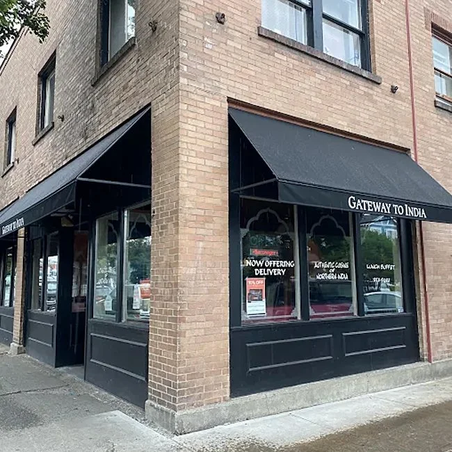 Corner of a brick building with black awnings displaying the sign 'Gateway to India'. The storefront has large windows with posters and signs, indicating food delivery services. Sidewalk in front of the store.