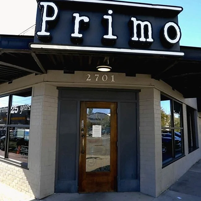 Exterior of a restaurant or bar called Primo with a sign displaying the word 'Primo' and the street number 2701 above the door. The building has large windows and a wooden door.
