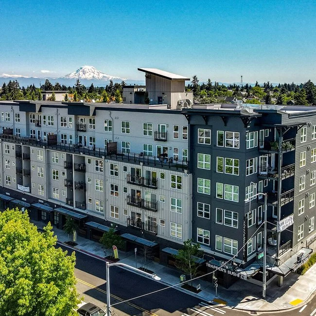 Modern multi-story apartment building with black and white facades, balconies, and a sushi restaurant on the ground floor, with a view of Mount Rainier in the background.
