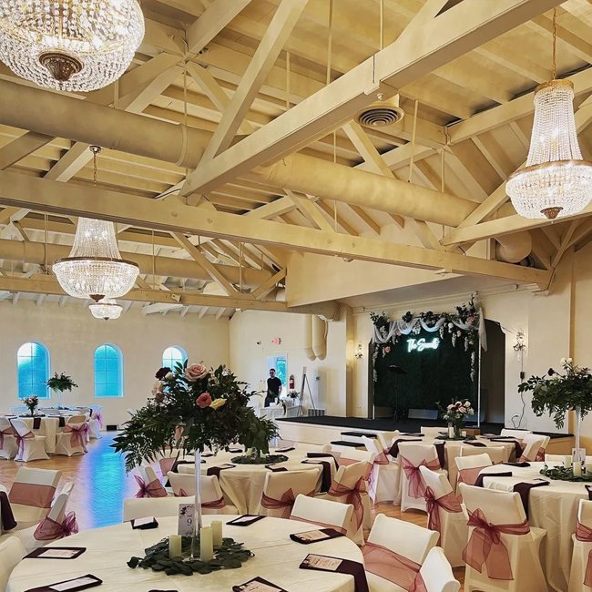 Elegant event hall with round tables decorated with pink and cream tablecloths and bows, floral centerpieces, and a stage with draped fabric and floral accents, illuminated by large chandeliers and wall-mounted lights.