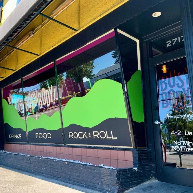 Storefront window with colorful mountain landscape mural, advertisements for drinks, food, rock & roll, and a partially visible sign for Bus Buddy.