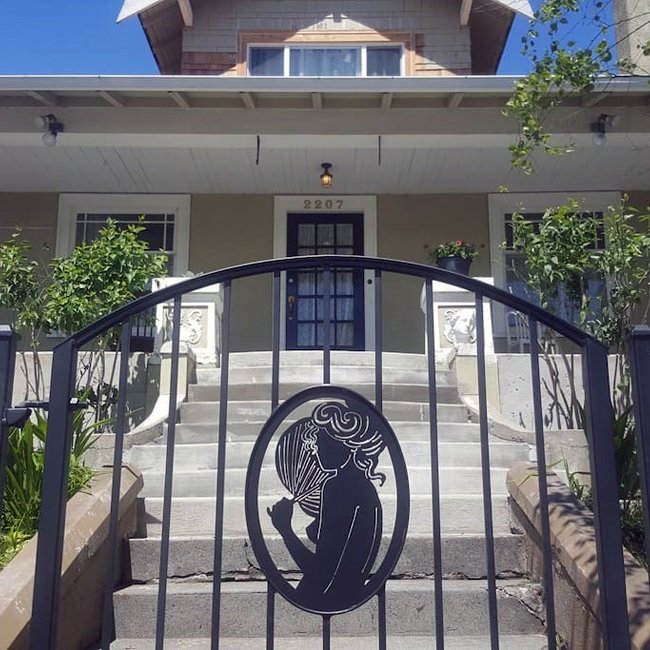 Front view of a house with stairs leading to a door, a decorative gate with a silhouette of a woman with flowing hair, and greenery on either side of the stairs.