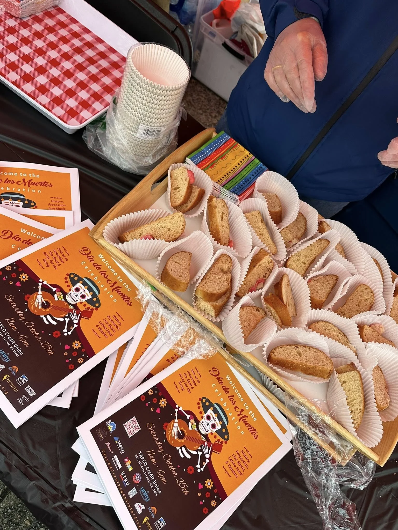 A person in blue clothing stands near a table with decorated flyers, paper cups, and a wooden tray filled with sliced cake or bread in paper liners. The flyers welcome visitors to a Día de los Muertos celebration.