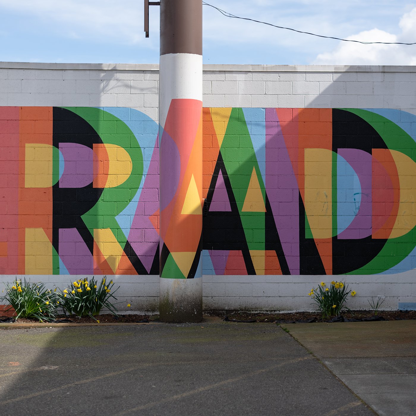 Colorful street art mural on a brick wall with a large flower in front, featuring bright overlapping geometric shapes in various colors and patterns.