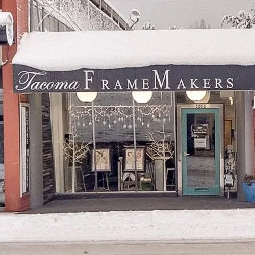 Exterior view of Tacoma Frame Makers storefront with a black awning displaying the shop's name, large front windows, and a turquoise door.