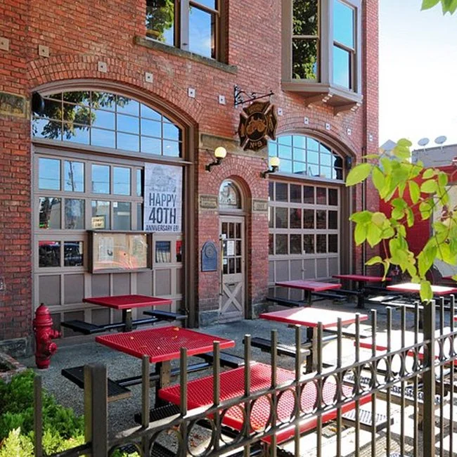 Exterior of a brick building with large windows, outdoor seating with red tables, a black metal fence, and a sign celebrating a 40th anniversary. There's a hanging sign with a dog logo and outdoor lights.