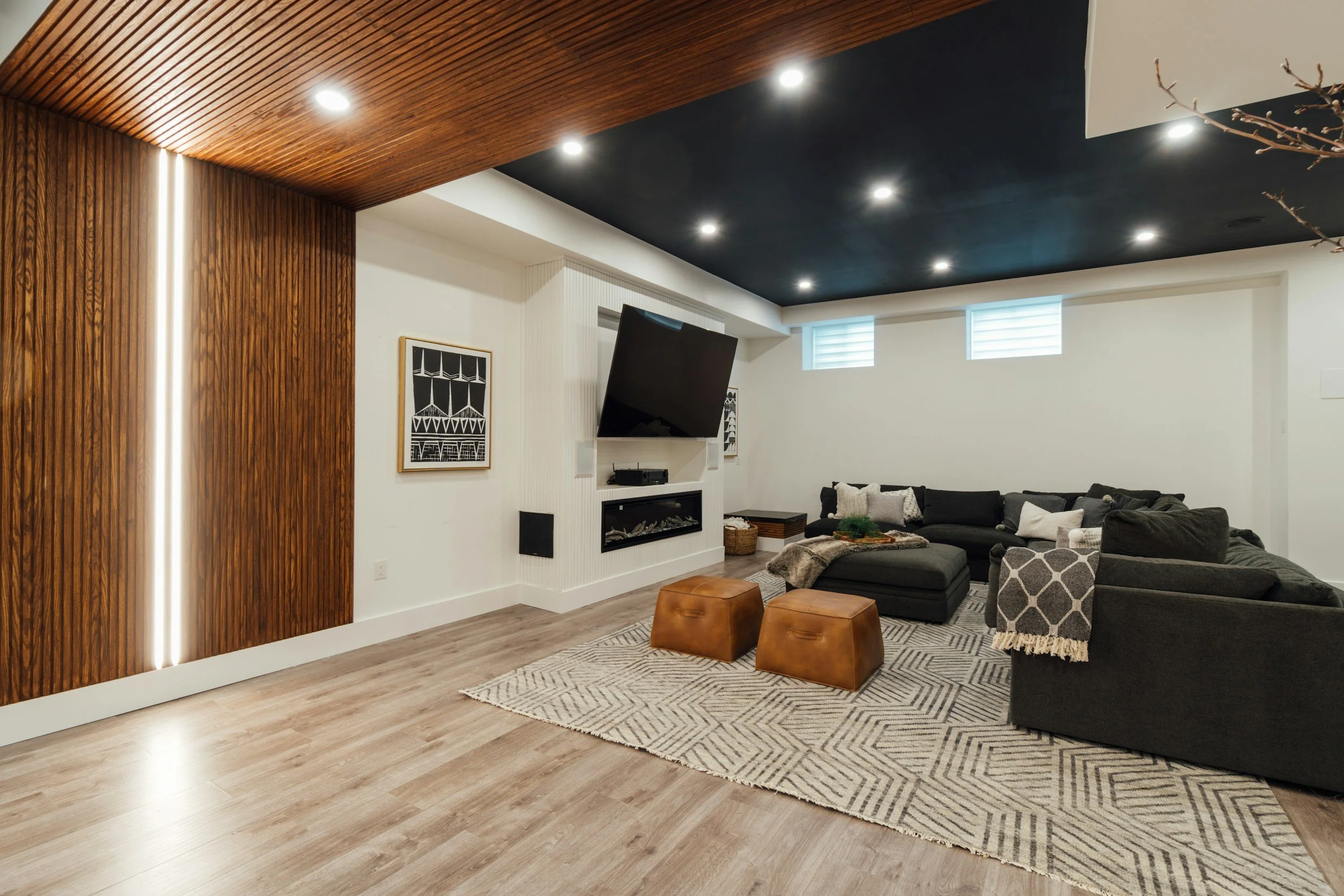 Modern living room with wood-paneled wall, black ceiling, and recessed lighting. Features a large black sectional sofa with throw pillows, a geometric area rug, and two brown leather ottomans. Mounted flat-screen TV above a fireplace with decorative logs.
