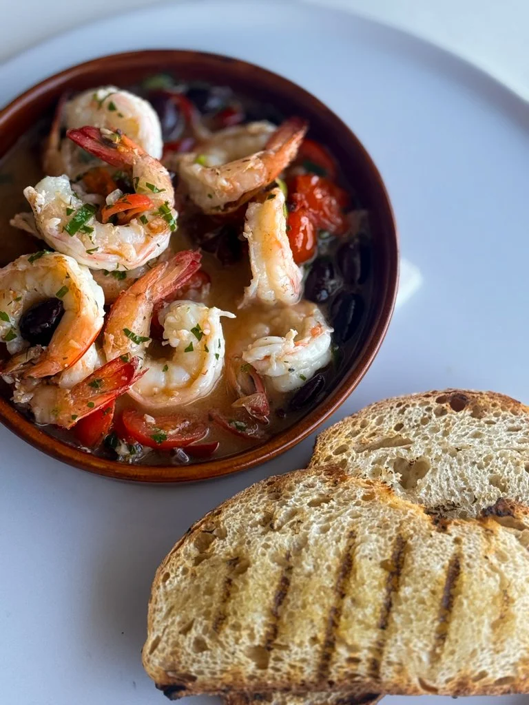 Sizzling Mooloolaba Prawns with garlic, herbs, chilli, white wine, kalamata olives & sourdough in a brown bowl.