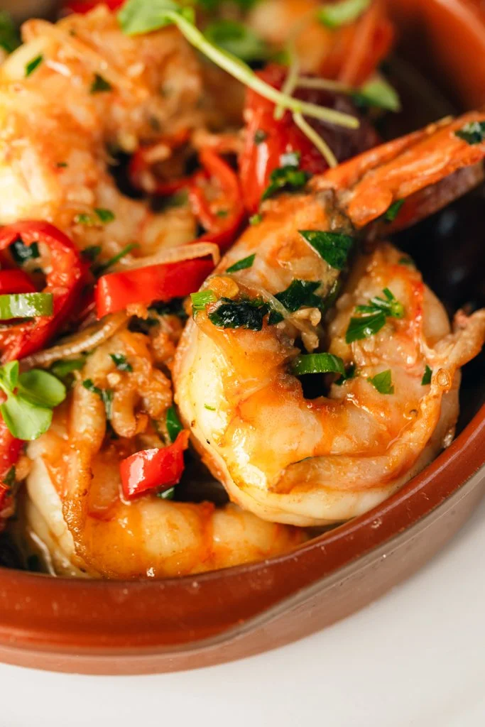 Sizzling Mooloolaba Prawns with garlic, herbs, chilli, white wine, kalamata olives & sourdough in a brown bowl.